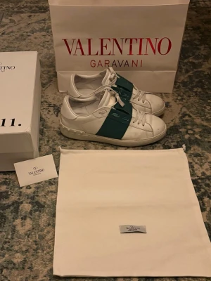 Valentino Garavani vita sneakers - Snygga vita sneakers från Valentino Garavani med en grön bred rem över sidan och klassiska nitar på hälen. Skorna har snörning och är tillverkade i skinn för en exklusiv känsla. Perfekta för dig som vill ha en stilren men ändå unik look.