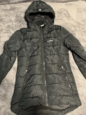 Svart pufferjacka från Everest - Snygg svart pufferjacka från Everest med huva och dragkedja framtill. Jackan har två dragkedjeförsedda fickor och quiltad design. Perfekt för kyliga dagar och har en längre passform som ger extra skydd mot vind.