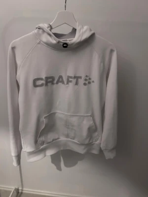 Vit hoodie från Craft med logga - Snygg vit hoodie från Craft med stor grå logga broderad på bröstet. Hoodien har en klassisk känguruficka framtill och en rymlig huva med meshfoder. Perfekt för chill dagar eller träning. Mjuk och bekväm bomullsblandning.