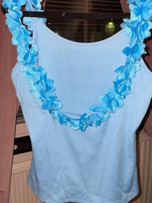 Ljusblå ribbad topp med blommor - Supersöt ljusblå ribbad topp med breda axelband och dekorativa blå blommor längs urringningen. Perfekt för dig som vill sticka ut med en unik och lekfull stil. Materialet känns mjukt och stretchigt, och toppen är ärmlös med en figurnära passform.
