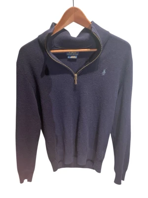 Mörkblå halvzip-tröja från Polo Ralph Lauren - Mörkblå stickad tröja från Polo Ralph Lauren med halv dragkedja och broderad logga på bröstet. Tröjan har ribbade muddar vid ärmslut och nederkant samt hög krage. Perfekt för lager på lager och klassisk stil.