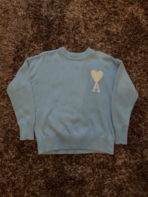 Ljusblå stickad tröja Ami Paris - Ljusblå stickad tröja från Ami Paris med rund halsringning och ribbade muddar. På bröstet finns ett vitt hjärta med bokstaven 'A' under, vilket är Ami Paris ikoniska logga. Tröjan har en avslappnad passform och är perfekt för lager på lager.