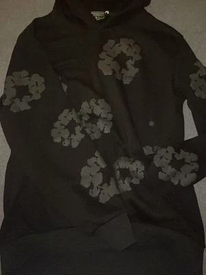 Svart hoodie med blommönster - Snygg svart hoodie från Marimekko med stora, gråa blommönster över hela tröjan. Klassisk huva med dragsko och ribbade muddar vid ärmslut och nederkant. Perfekt för dig som vill sticka ut med ett unikt tryck och samtidigt hålla det stilrent. Säljer eftersom den är för stor för mig och den passar ej mig.