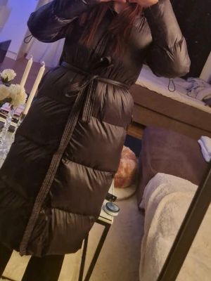 Svart lång dunjacka med bälte - Snygg svart lång dunjacka med knytbälte i midjan och quiltad design. Jackan har lång ärm och en klassisk pufferstil som håller dig varm under kalla dagar. Perfekt för dig som vill ha både stil och funktion.