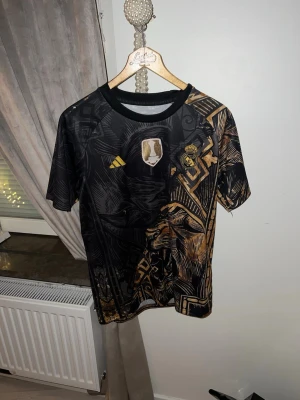 Fotbollströja  - Snygg svart Real Madrid matchtröja från Adidas med guldiga detaljer och ett stort lejonmotiv. Tröjan har klubbmärke och Adidas-logga på bröstet samt unikt grafiskt mönster. Tillverkad i lätt och ventilerande material, perfekt för träning 