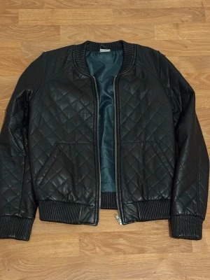 Svart quiltad bomberjacka i skinnimitation - Snygg svart bomberjacka i skinnimitation med quiltat mönster. Jackan har ribbade muddar vid ärmslut, hals och nederkant samt dragkedja framtill. Perfekt för dig som vill ha en stilren och trendig look. Vid axeln har den en liten skada som syns inte mycket 