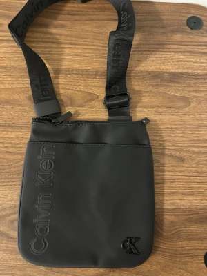 Svart axelväska från Calvin Klein - Snygg svart axelväska från Calvin Klein med justerbart axelband och diskret logga framtill. Väskan har en stilren design med dragkedja upptill och är tillverkad i slitstarkt syntetmaterial. Perfekt för att bära dina viktigaste grejer.