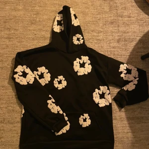 Svart hoodie med vita blommor - Snygg svart hoodie med stora vita blommönster över hela tröjan och huvan. Klassisk känguruficka framtill och ribbade muddar vid ärmslut och nederkant. Perfekt för dig som gillar streetwear och vill sticka ut med ett unikt mönster.