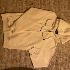 Vit zip hoodie från Polo Ralph Lauren - Säljer en stilren vit hoodie med dragkedja från Polo Ralph Lauren. Hoodien har huva med dragsko, två fickor framtill och den klassiska lilla loggan broderad på bröstet. Perfekt för en clean och avslappnad look.
