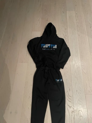 Svarta mjukisbyxor från Trapstar - Svarta mjukisbyxor från Trapstar med matchande hoodie. Byxorna har resår i midjan och tryckt Trapstar-logga i blått och vitt på benet. Materialet är mjukt och skönt, perfekt för chill dagar. Snygg streetwear-vibe med trycket 'It's a secret' på både byxa och hoodie.