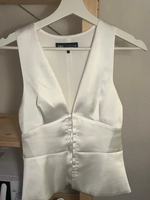 Vit satinlinnetopp från Zara - Säljer en elegant vit linnetopp från Zara i glansigt satinmaterial. Toppen har djup v-ringning, breda axelband och figurnära passform. Perfekt att styla med jeans eller kjol för en trendig look.
