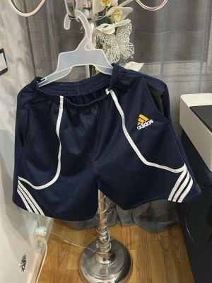Marinblå Adidas träningsshorts - Säljer ett par marinblå träningsshorts från Adidas med vita detaljer och klassiska ränder. Shortsen har elastisk midja och Adidas-logga broderad i gult och vitt på ena benet. Perfekta för sport eller chill dagar.