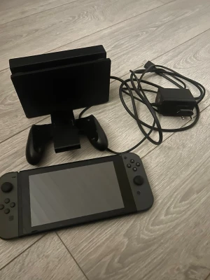 Nintendo Switch med Joy-Con och docka - Nintendo Switch spelkonsol i svart med tillhörande blå och röd Joy-Con, dockningsstation, handkontrollshållare och laddare. Konsolen har en rektangulär form och är tillverkad i plast. Perfekt för gaming både hemma och på språng.