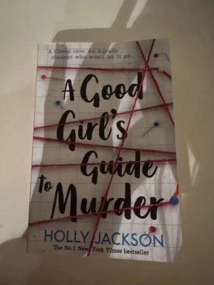 A Good Girl's Guide to Murder - En spännande pocketbok med titeln 'A Good Girl's Guide to Murder' av Holly Jackson. Omslaget är vitt med rutmönster, röda trådar och nålar som ger en mystisk känsla. Perfekt för dig som gillar deckare och thrillers.
