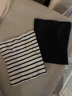 Två bandeau tubtoppar, svart & randig - Säljer två bandeau tubtoppar, en i helsvart och en i vitt med svarta ränder. Perfekta till sommaren och lätta att matcha med olika outfits. Båda är ärmlösa och har en enkel, stilren design.