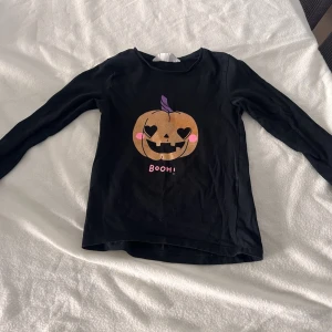 Svart halloweentröja med pumpatryck - Svart långärmad tröja med ett tryck av en pumpa med hjärtformade ögon, lila stjälk och texten 'BOOH!' i rosa. Perfekt för halloween eller maskerad. Mjuk och bekväm bomullskänsla.