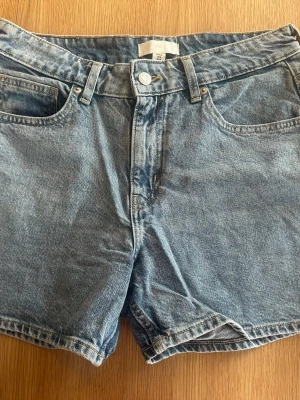 Blå denimshorts från hm - Klassiska blå denimshorts från hm med fem fickor, bälteshällor och knappgylf. Shortsen har en rak passform och är tillverkade i bomull. Perfekta för sommaren och enkla att matcha med olika stilar.