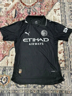 Manchester City svart Puma Third kit matchtröja - Svart Manchester City fotbollströja Spelarversion från Puma med klassisk krage och vita detaljer. Tröjan har korta ärmar, broderad klubbmärke och Puma-logga på bröstet samt Etihad Airways-tryck. Tillverkad i lätt funktionsmaterial (Ultraweave) för maximal komfort.