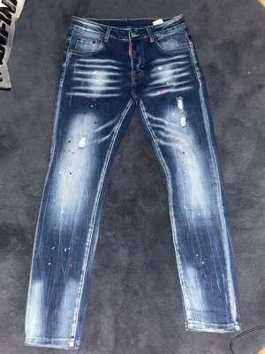 Dsquared2 blå jeans med patch - Säljer ett par blå jeans från Dsquared2 med slitningar, färgstänk och en stor röd och gul patch med text och siffran 2 på bakfickan. Jeansen har klassisk femficksmodell, knappgylf och smal passform. Perfekt för dig som vill sticka ut. Köpte fel storlek