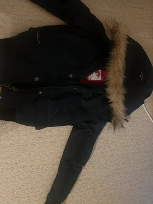 Svart parkas med fuskpäls från Canada Goose - Snygg svart parkas från Canada med huva och fuskpälsdetalj. Jackan har flera fickor, dragkedja och knappar framtill samt ribbade muddar vid ärmsluten. Perfekt för kalla dagar och har en klassisk, tidlös look. Passar perfekt till barn och är jätte varm. DM för mer bilder. Orignal äkta jacka, användts endast två tre gånger