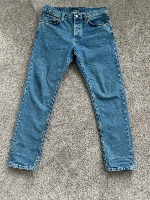 Blå straight jeans från Nudie Jeans - Klassiska blå jeans från Nudie Jeans med rak passform och fem fickor. Jeansen har normal midja och är tillverkade i slitstark bomullsdenim. Snyggt broderade bakfickor och knappgylf framtill. Vid frågor är det bara att slänga iväg ett meddelande!