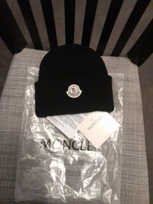 Svart  mössa i ull - Snygg svart ribbstickad mössa från Monscler med uppvikt kant och klassisk Monscler-logga framtill. Tillverkad i mjuk ull som håller dig varm och stilren under kalla dagar. Perfekt accessoar för dig som vill ha både komfort och trendig look.