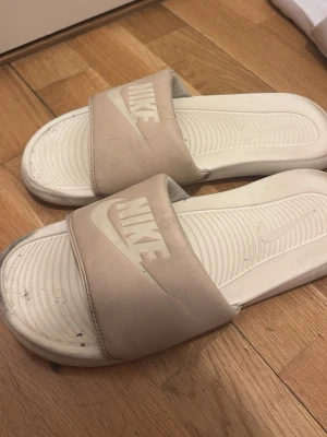 Vita Nike tofflor med logga - Två par klassiska Nike tofflor i vitt med bred rem över foten och tydlig Nike-logga. Ena paret har beige ton på remmen och det andra paret är helvita med svart logga. Snygg och enkel design, perfekt för chill dagar eller efter träning.
