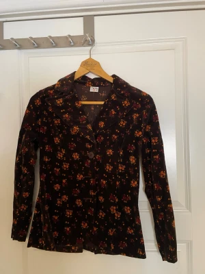 Blommig brun kavaj och kjol från PETRI - Snygg brun kavaj och kjol från PETRI med blommigt mönster i gult och rött. Kavajen har klassisk krage, långa ärmar och knäppning framtill.