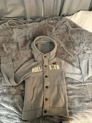Grå cardigan från Hollister med knappar - Snygg grå cardigan från Hollister med knäppning framtill och broderad logga i vitt och rött på bröstet. Tröjan har huva med dragsko, två fickor och ribbade muddar. Perfekt för en avslappnad och cool stil. En liten defekt (sista bilden)