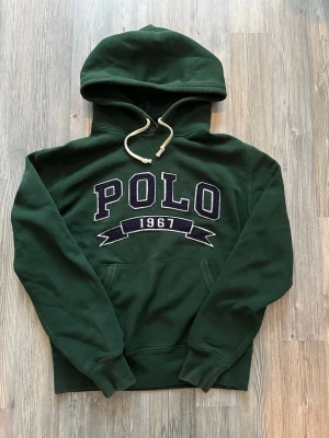 Grön hoodie från Polo Ralph Lauren - Säljer nu en Mörkgrön hoodie från Polo Ralph Lauren med stor POLO-logga och '1967’ broderat på magen. Storleken är runt S och bör passa någon mellan 170-180. Har en liten liten fläck på ett av snörena men det märkte jag inte förens jag skulle ta kort på tröjan så det är inget som någon kommer märka. Skriv gärna vid frågor eller funderingar😇🙏