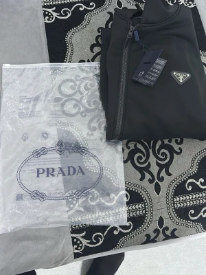Svart hoodie med dragkedja från Prada - Säljer en svart hoodie från Prada med dragkedja framtill och klassisk huva med dragsko. Hoodien har fickor på magen och den ikoniska Prada-loggan på bröstet. Materialet känns mjukt och exklusivt, perfekt för dig som vill ha en stilren och lyxig look.