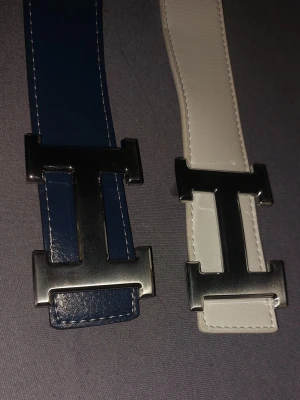 Två bälten - Säljer två stilrena bälten med stort H-format metallspänne. Ett bälte är mörkblått och det andra är vitt, båda i slätt läder med synliga sömmar. Perfekta accessoarer för att lyfta din outfit.