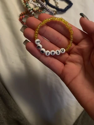 Gult pärlarmband med texten ZENITSU - Handgjort armband med små gula pärlor och vita runda bokstavspärlor som bildar namnet ZENITSU. Elastisk tråd gör det enkelt att ta på och av. Perfekt accessoar för dig som gillar anime eller vill ha ett personligt smycke.