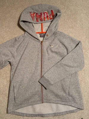 Grå Puma ziphoodie med roséfärgad dragkedja, corall färgat märke. - Snygg grå hoodie från Puma med roséfärgad dragkedja framtill och huva med corall/aprikos PUMA-logga på huvan. Klassisk passform, långa ärmar och diskret Puma logga på bröstet. Mjuk flossning invändigt.