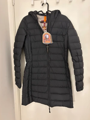 Parajumpers Irene jacka  - Snygg svart lång dunjacka från Parajumpers med huva och dragkedja framtill. Jackan har quiltad design och är perfekt för kalla dagar. Klassisk logga på ärmen och smickrande passform. Perfekt för dig som vill hålla stilen även när det är kallt ute. Modell Irene kvitto och prislappar finns 