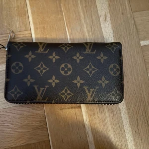 Plånbok med mönster - Helt ny plånbok med mönster. Även Mussepigg på en sida. Fack för kort, sedlar och fack för mynt med dragkedja. Följer med en sling, så man kan bära den på handleden också, glömde fota den, skriv om ni vill se den. Är en svart sling. Följer med dustbag och kartong till också. Kan skicka bild på dom också. Lapp sitter kvar också. Är ca 19-20cm bred och va 11-12cm hög