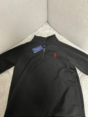 Svart half-zip tröja Polo Ralph Lauren - Svart slim fit tröja från Polo Ralph Lauren med half-zip och röd broderad logga på bröstet. Tillverkad i stretchmesh-material som ger en skön och flexibel passform. Perfekt för en clean och stilren look.