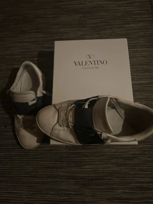 Valentino skor - Snygga vita sneakers från Valentino Garavani med bred blå detalj över vristen och klassisk snörning. Skorna har rund tå och platt sula, tillverkade i skinn för en exklusiv känsla. Perfekta för dig som vill ha en stilren och trendig look. Är strl 43/44 skriv för mer frågor!