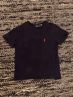 Marin blå t-shirt från Polo Ralph Lauren - Klassisk marin blå t-shirt från Polo Ralph Lauren med röd broderad logga på bröstet. T-shirten har rund halsringning och korta ärmar. Tillverkad i mjuk bomull för en skön känsla och enkel stil.