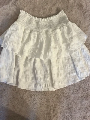 Vit volangkjol från SHEIN - Supersöt vit kortkjol från SHEIN med dubbla volanger och smock i midjan. Kjolen har en luftig och lätt struktur, perfekt för varma dagar. Passar dig som gillar en feminin och trendig stil.