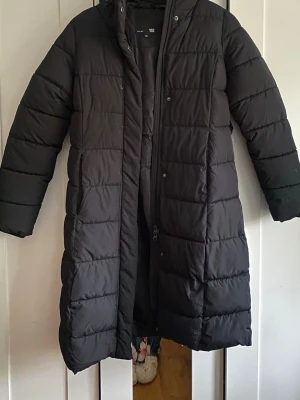 Svart lång dunjacka med huva - Snygg svart lång dunjacka med huva och quiltad design. Jackan har dragkedja och tryckknappar framtill samt långa ärmar. Perfekt för kalla dagar när du vill hålla dig varm och stilren.