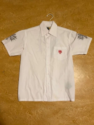 Vit skjorta med tribaltryck och brodyr - Kortärmad vit skjorta med tribaltryck i grått på axlarna och röd broderad detalj på bröstfickan. Klassisk krage och knäppning framtill. Skjortan är i bomull och har en modern, edgy vibe. Står S men passar även M checka måtten. Mått: längd: 68cm bredd: 51cm 