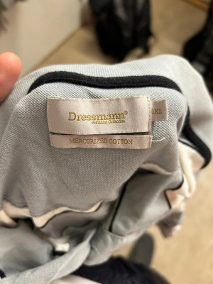 Randig pikétröja från Dressmann - Klassisk pikétröja från Dressmann i merceriserad bomull. Tröjan har breda horisontella ränder i ljusgrått och vitt, korta ärmar och krage med knappar. Perfekt för en avslappnad och stilren look.