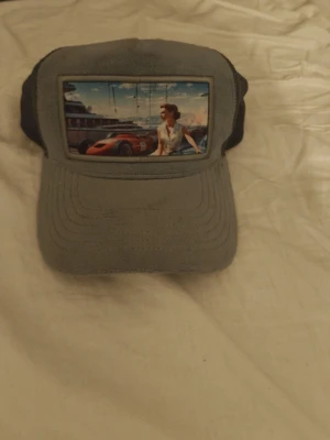 Grå meshkeps med retrotryck - Säljer en grå keps med mesh på sidorna och ett stort retroinspirerat tryck framtill som visar en kvinna, en röd sportbil och en yacht i bakgrunden. Kepsen har böjd skärm och justerbar baksida. Perfekt för dig som gillar unik streetwear och coola motiv.