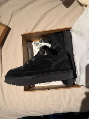 UGG lowmel - Snygga svarta sneakers från UGG med robust platt sula och rund tå. Ovandelen är i mocka och textil, och skorna har snörning samt fuskpäls runt öppningen för extra komfort och stil. Perfekta för dig som vill ha både värme och streetstyle.                                  Då de är för små säljer jag de. Helt nya aldrig använda och bara testade en gång de är en storlek för små så de här passar 44/43 men i beskrivningen är de 45:)