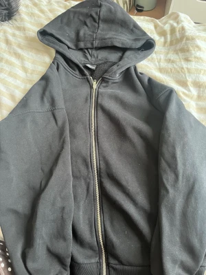 Svart zip hoodie från Gina Tricot - Klassisk svart hoodie från Gina Tricot med dragkedja framtill och stor huva. Tillverkad i mjukt och lite tjockare material med borstad insida för extra komfort. Perfekt för en avslappnad och stilren look.