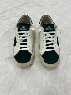 The Gallerist vita och gröna sneakers - Snygga sneakers från The Gallerist i vitt och mörkgrönt med coolt klubbmärke på sidan. Ovandel i skinn och mocka, vita snören och platt sula. Tungan har broderad logga och insidan är mjukt fodrad. Perfekta för dig som gillar unik stil och detaljer. Storlek 41 men en person med 43 kan ha på sig dem