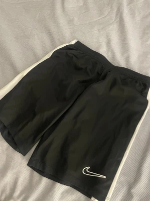 Svarta Nike Dri-Fit shorts L - Säljer ett par svarta Nike Dri-Fit shorts med vita sidor och klassisk swoosh-logga framtill. De är tillverkade i ett lätt och snabbtorkande material som passar perfekt för träning. Elastisk midja för skön passform.