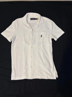 Polo Ralph Lauren kortärmad skjorta - Vit kortärmad skjorta från Polo Ralph Lauren i mjuk frotté. Klassisk krage, knappstängning framtill och bröstficka med mörkblå broderad logga. Perfekt för chill sommardagar när du vill ha en clean och avslappnad look.