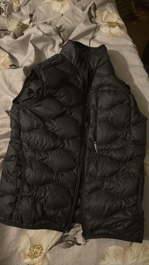 Svart dunväst Peak Performance S - Svart dunväst från Peak Performance i storlek S. Västen har ett quiltat mönster, hög krage och dragkedja framtill. Fickor med dragkedja på sidorna och diskret logga på insidan. Materialet är syntet och dun, perfekt för kyliga dagar.